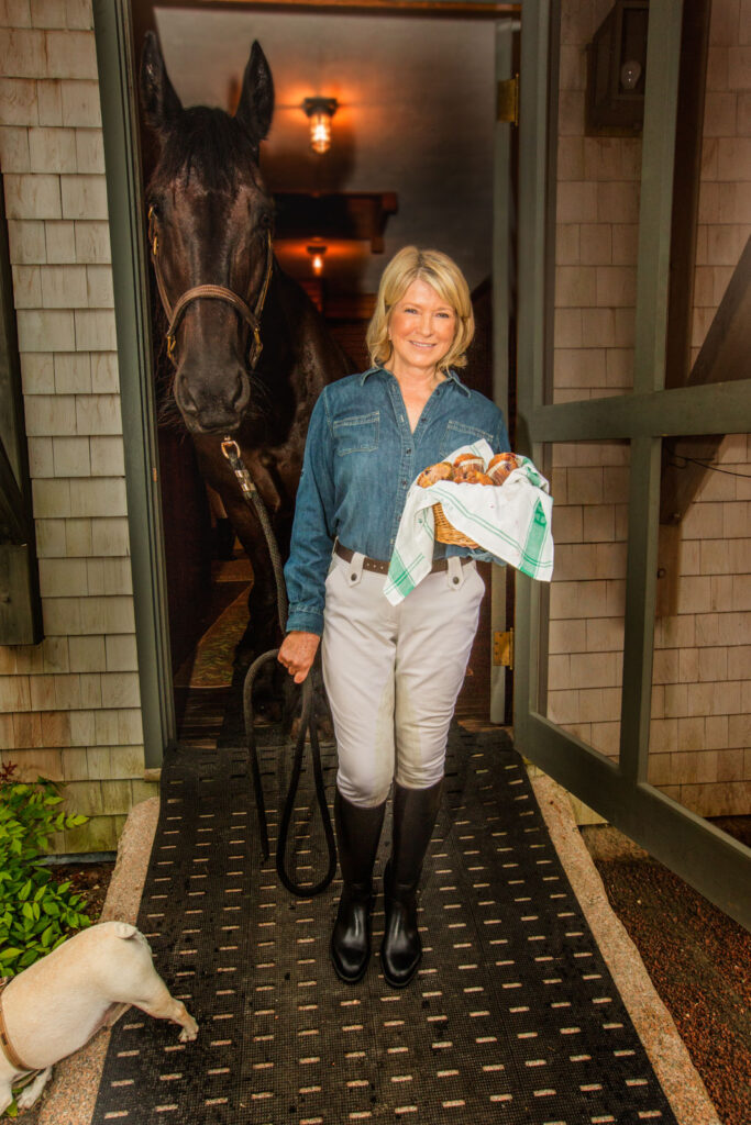 Martha Stewart holding a basket of muffins beside a Friesian horse in a barn doorway