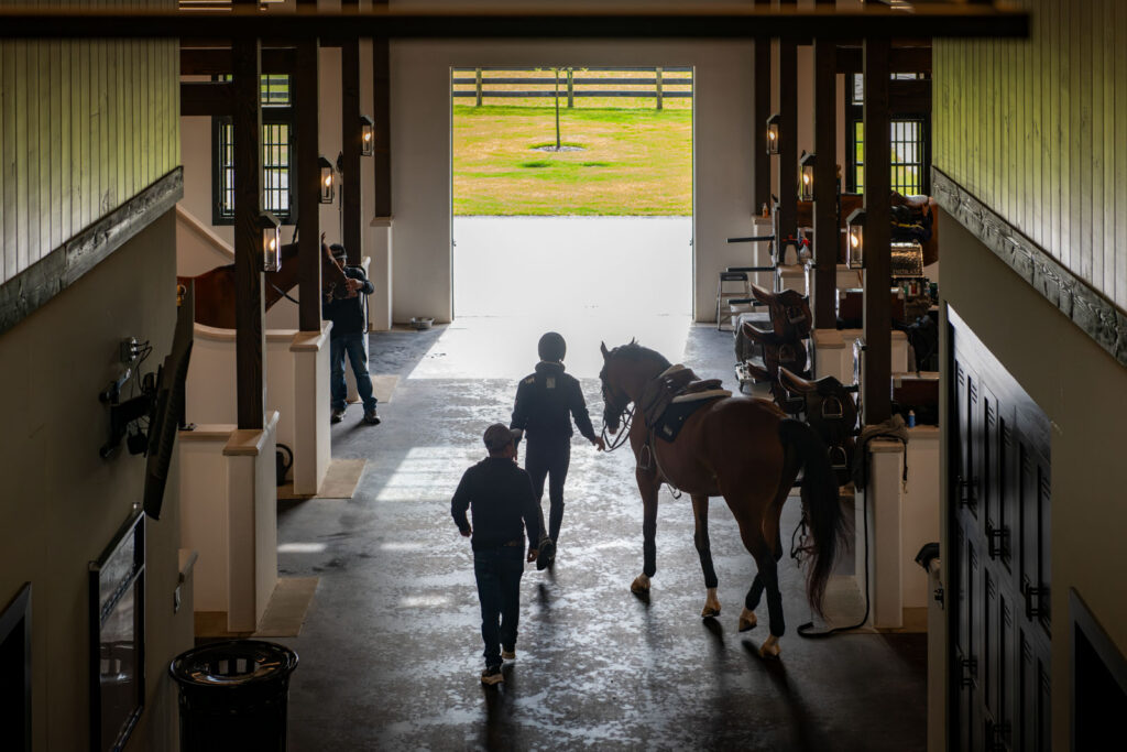 Overhead view of a groom leading a horse down the barn aisle toward the open door and the paddock beyond
