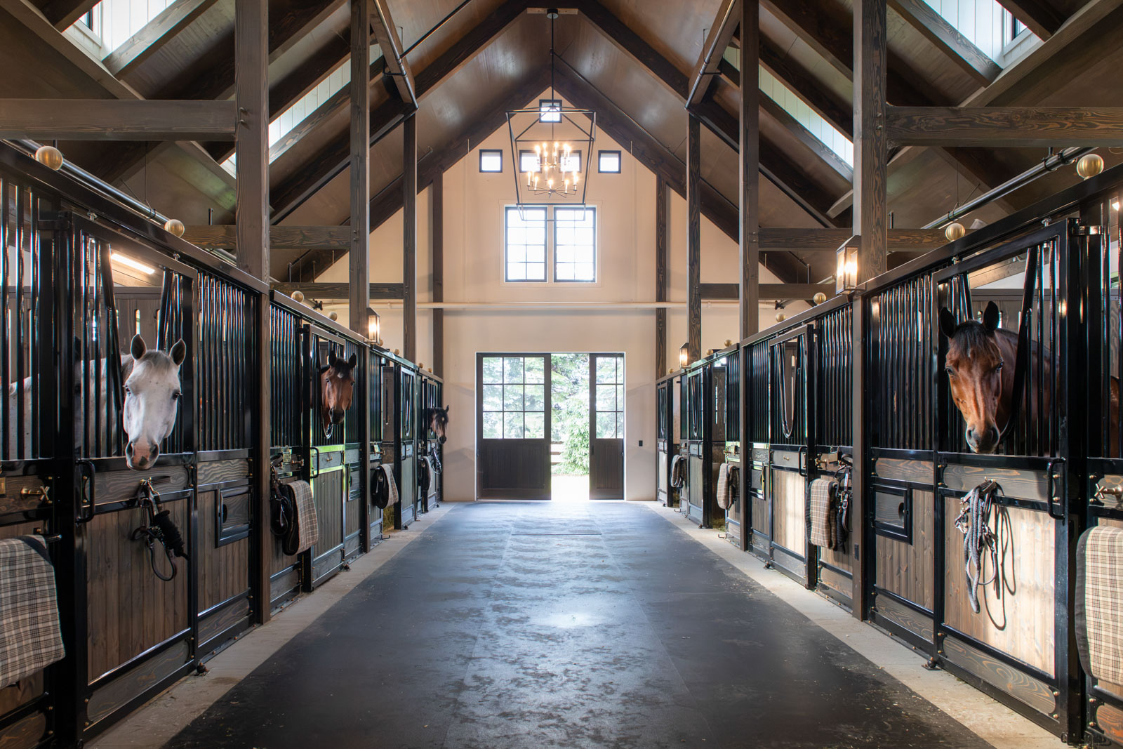 Timber-frame barn aisle with a lantern chandelier and horses looking out of black stall fronts at Chickering Traverse City