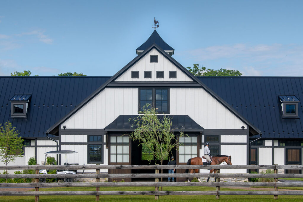 White board-and-batten barn with a black standing-seam metal roof and cupola behind a post-and-rail fence, with a rider passing in front