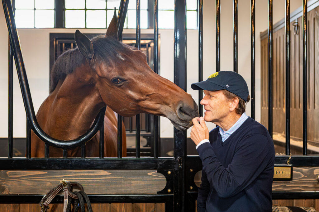 John Ingram in a navy sweater and cap, nose to nose with a bay horse over a black stall front